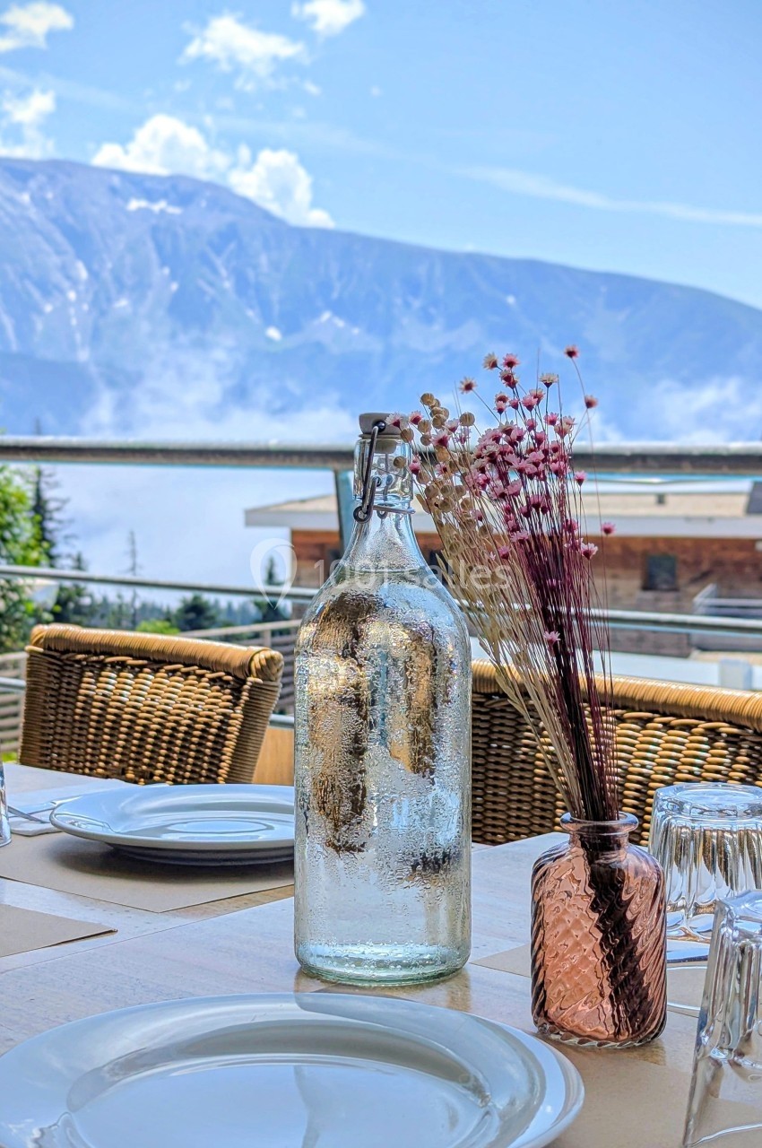 Bouteille en verre et vase avec fleurs séchées sur une table dressée en terrasse, avec vue sur des montagnes.