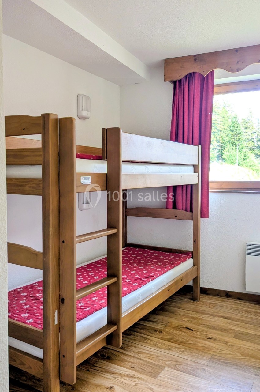 Lits superposés en bois avec matelas rouges dans une chambre lumineuse, parquet au sol et fenêtre avec rideaux rouges.