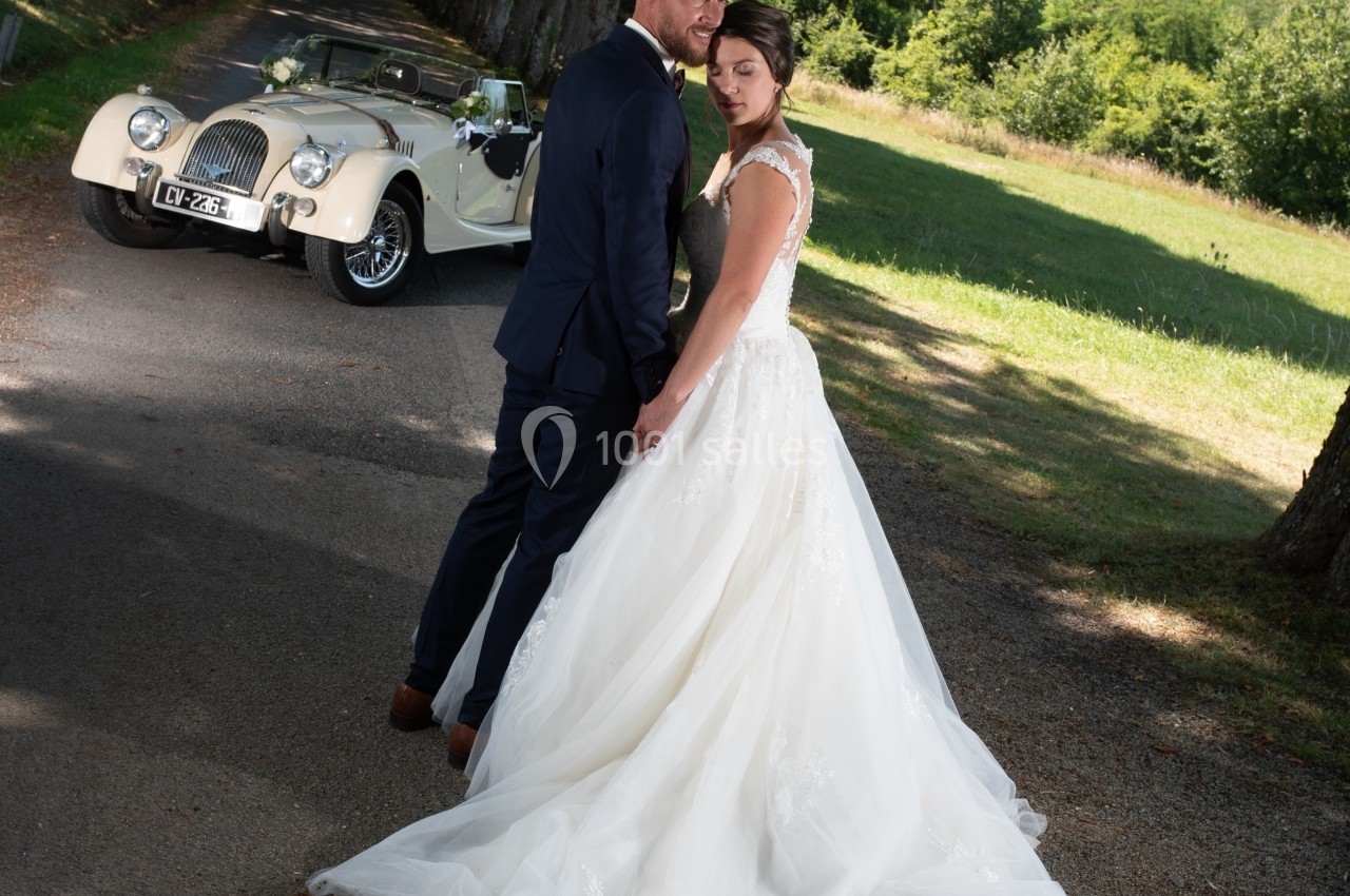 Un couple en tenue de mariage pose sur une allée bordée d'arbres, avec une voiture ancienne en arrière-plan.