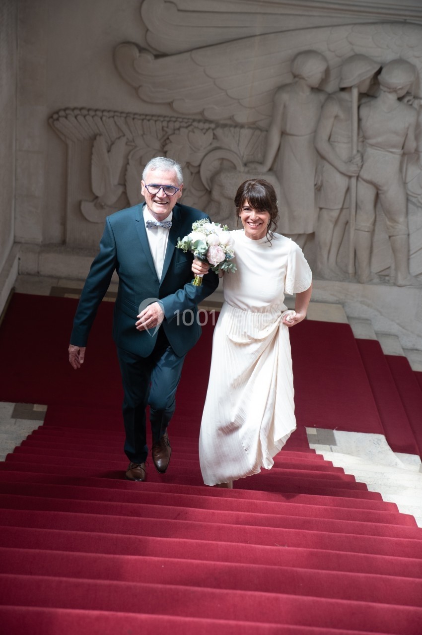 Un homme en costume et une femme en robe blanche montent un escalier rouge, souriants, avec un bouquet de fleurs.