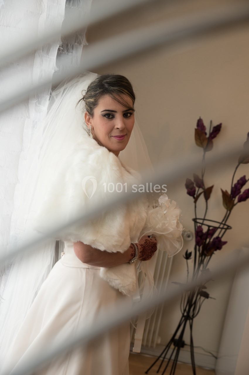 Femme en robe de mariée blanche, tenant un bouquet, posant près d'une décoration florale dans un intérieur lumineux.