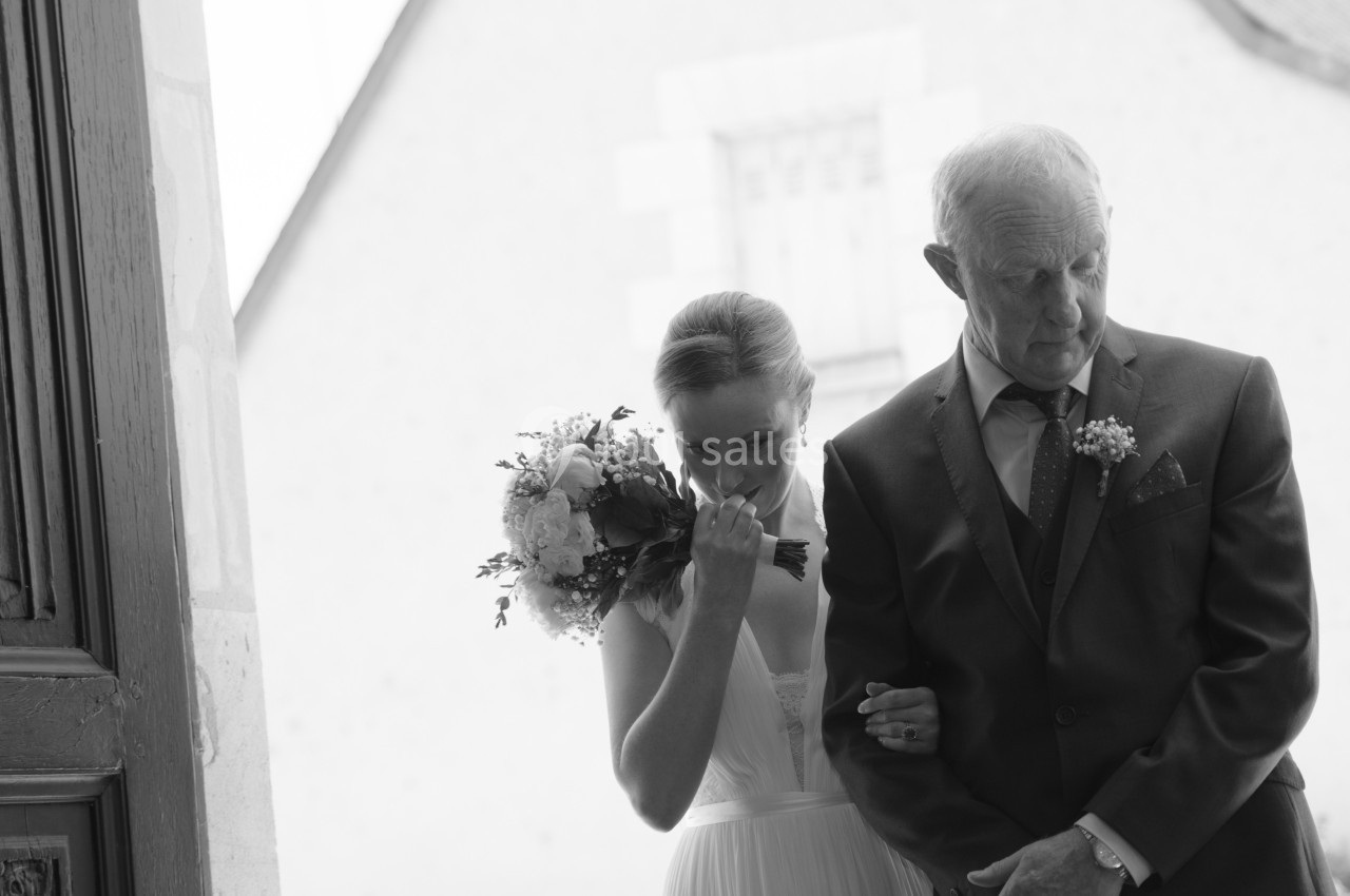 Une mariée en robe blanche entre dans un bâtiment au bras d'un homme en costume, tenant un bouquet de fleurs.