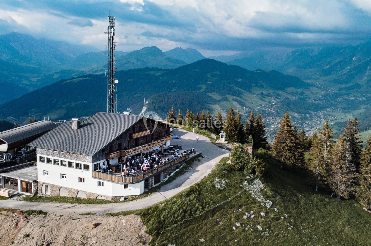 Vue aérienne d'un chalet en montagne entouré de sapins, avec une terrasse animée et un paysage vallonné en arrière-plan.