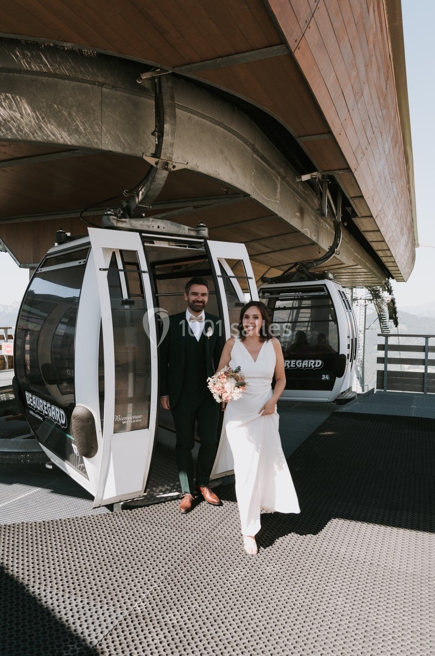 Un couple en tenue de mariage sort d'une cabine de téléphérique sur une plateforme ensoleillée.