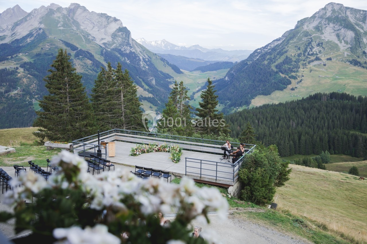 Terrasse en bois avec des chaises et des fleurs, offrant une vue panoramique sur des montagnes et une vallée verdoyante.