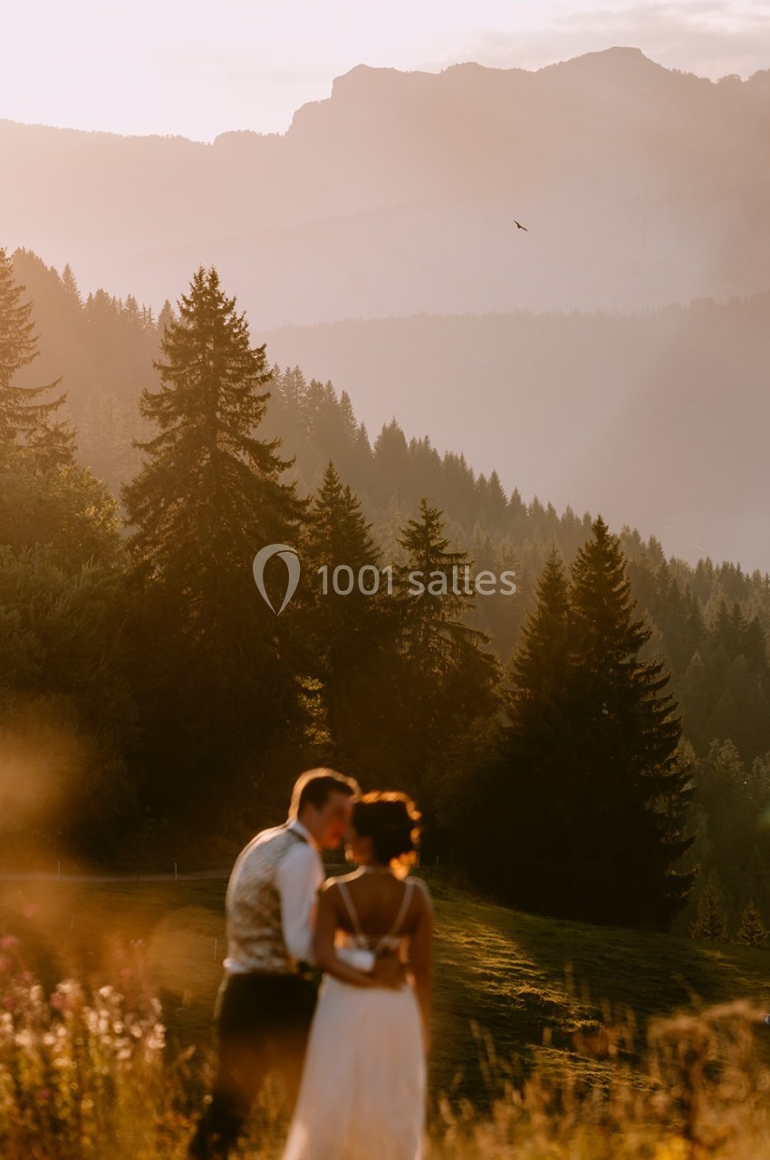 Un couple en tenue de mariage se tient dans un paysage montagneux au coucher du soleil, entouré de sapins.