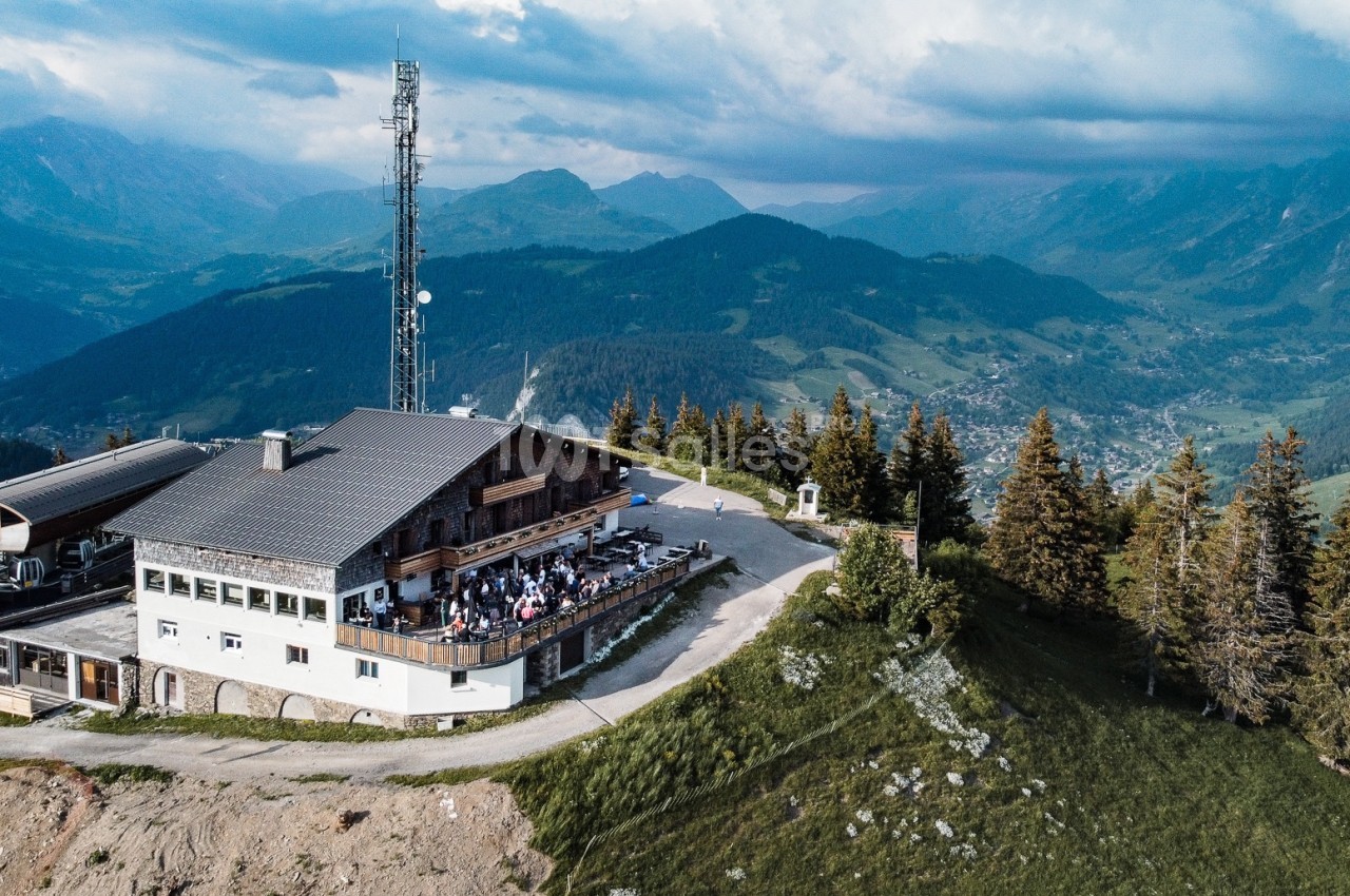 Vue aérienne d'un chalet en montagne avec une terrasse animée, entouré de forêts et de vallées verdoyantes.
