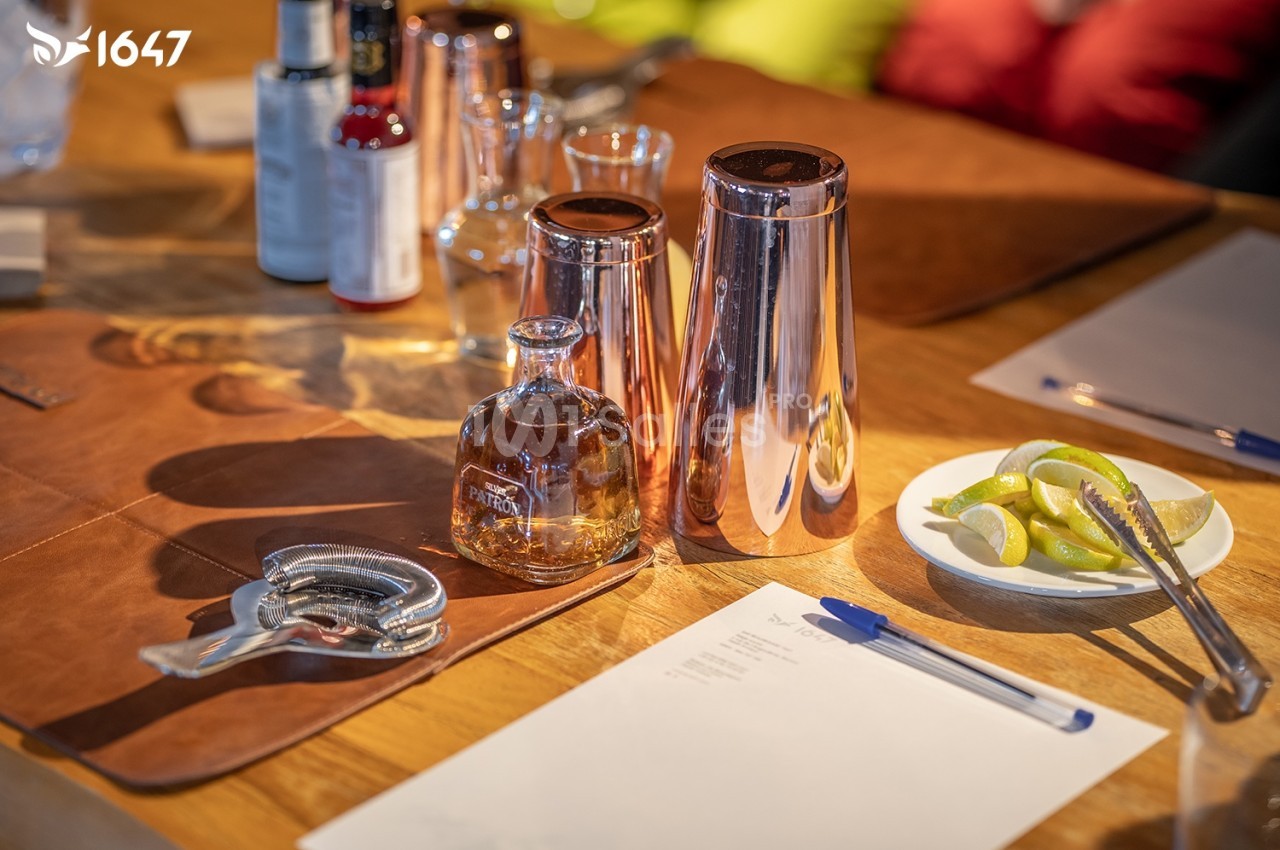 Ustensiles de bar, bouteille de spiritueux, assiette de quartiers de citron et feuilles de papier sur une table en bois.