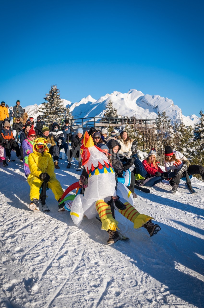 Des personnes déguisées descendent une piste enneigée en luge, entourées de spectateurs sous un ciel bleu.