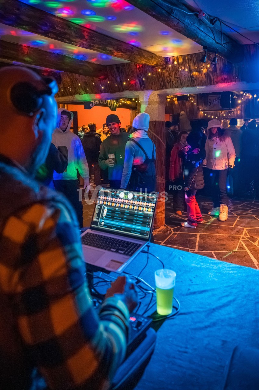 Un DJ mixe sur son ordinateur portable lors d'une soirée animée dans un espace intérieur éclairé par des lumières colorées.