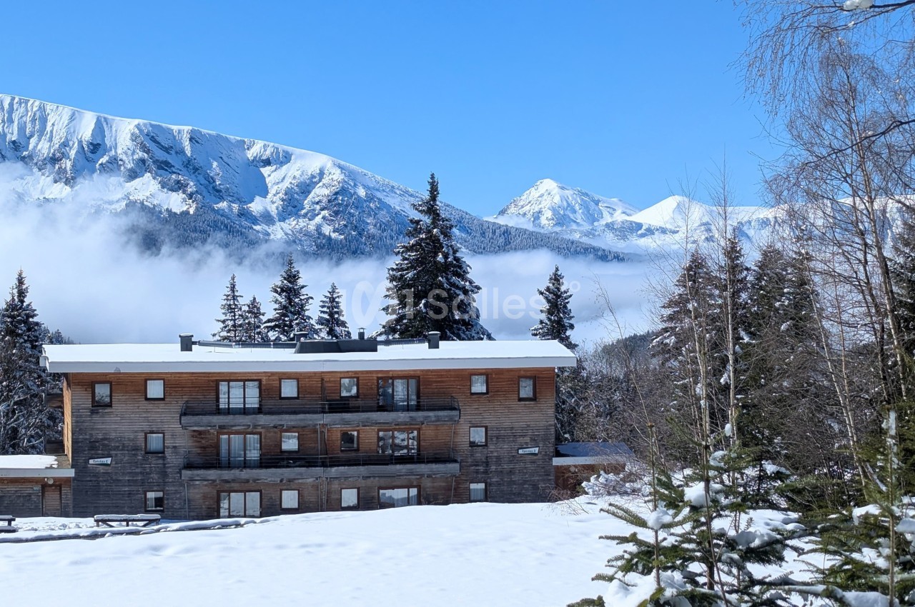 Chalet en bois entouré de neige, avec des montagnes enneigées et des sapins sous un ciel bleu en arrière-plan.