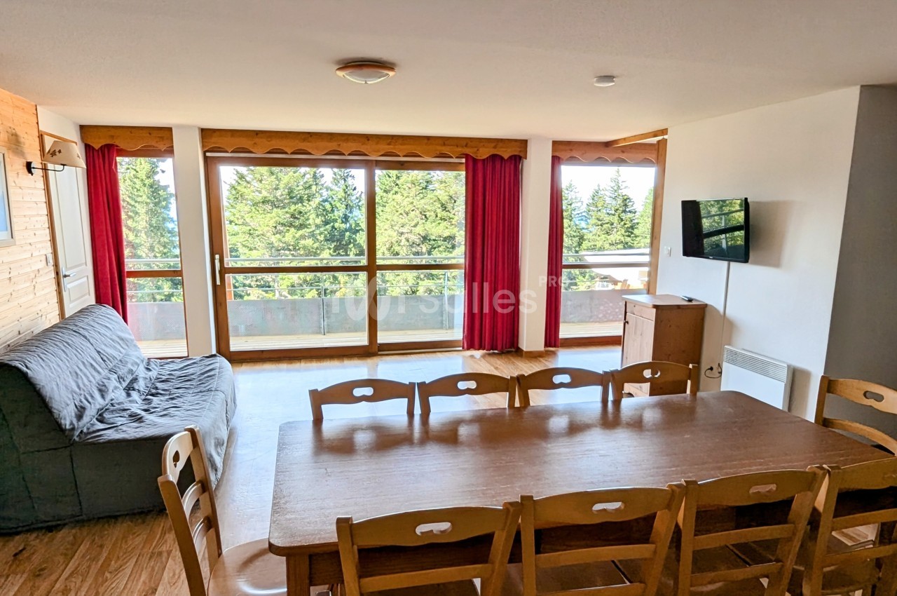 Salle à manger lumineuse avec table en bois, chaises, canapé, télévision murale et grandes baies vitrées donnant sur une…