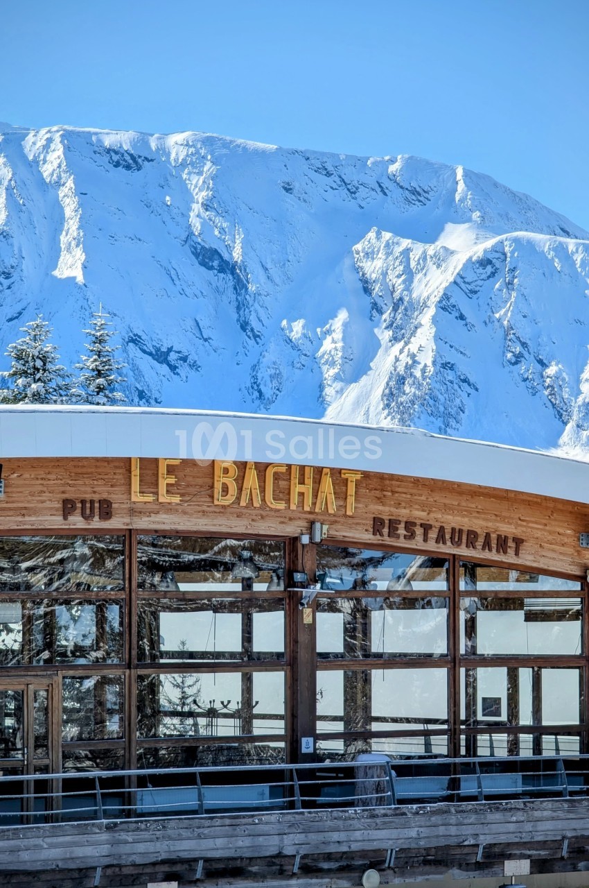 Façade en bois du restaurant ’Le Bachat’ avec des montagnes enneigées en arrière-plan sous un ciel bleu.