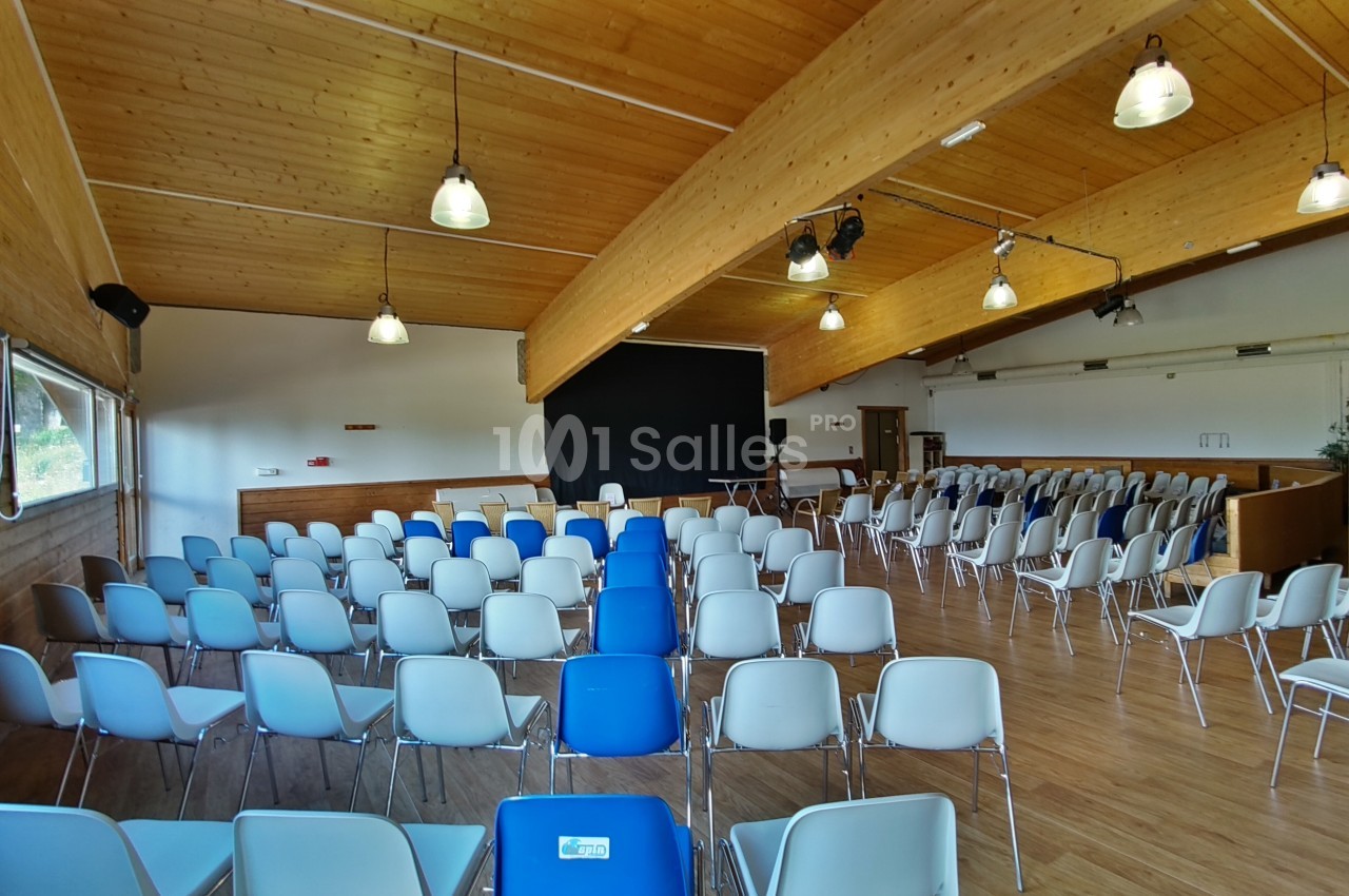 Salle de réunion avec des rangées de chaises blanches et bleues, un écran noir et un plafond en bois éclairé.