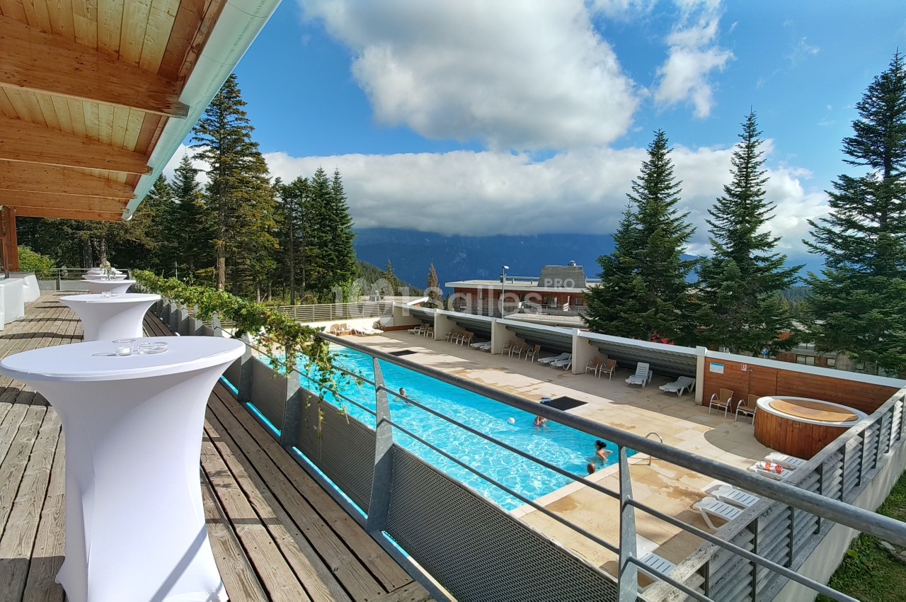 Terrasse en bois surplombant une piscine extérieure entourée de sapins, avec vue sur des montagnes sous un ciel…