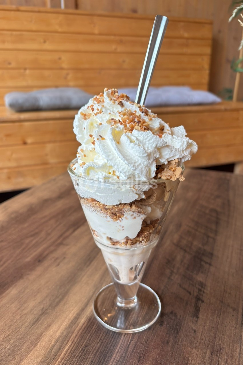 Coupe glacée avec chantilly, éclats de biscuits et cuillère en métal, posée sur une table en bois.