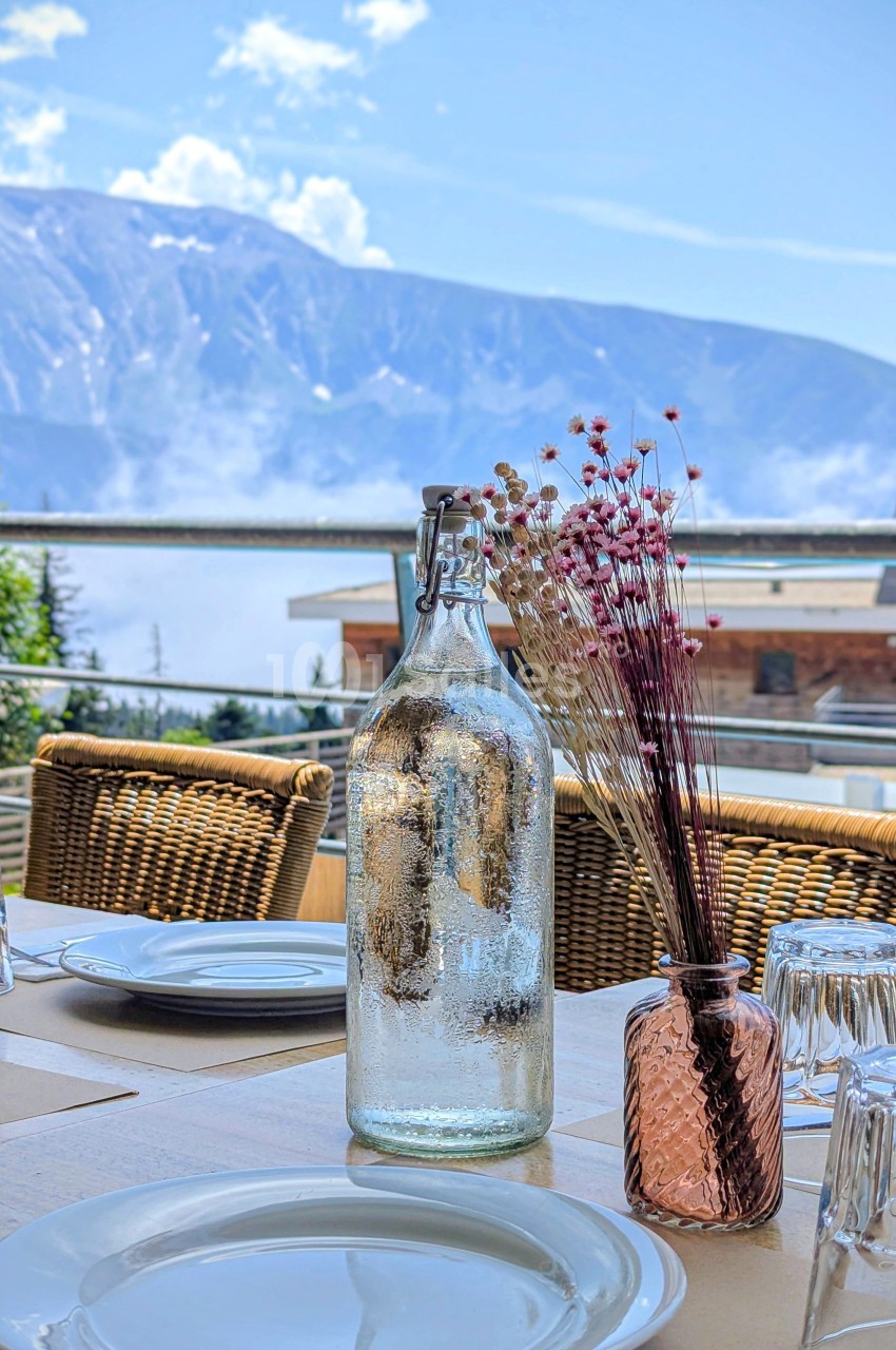 Bouteille en verre et vase avec fleurs séchées sur une table dressée en terrasse, avec vue sur des montagnes.