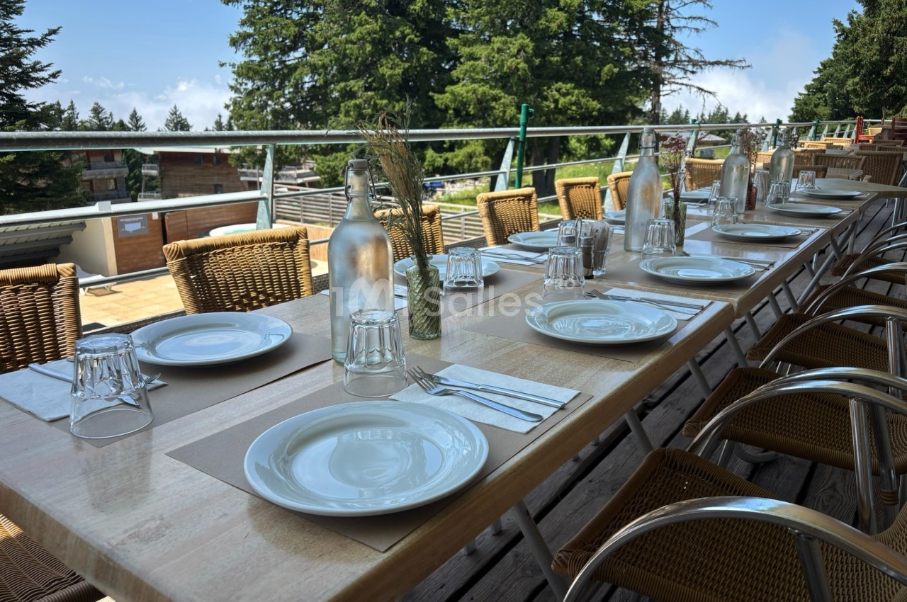 Table en bois dressée en extérieur avec assiettes, verres et carafes, entourée de chaises en osier, vue sur des arbres.