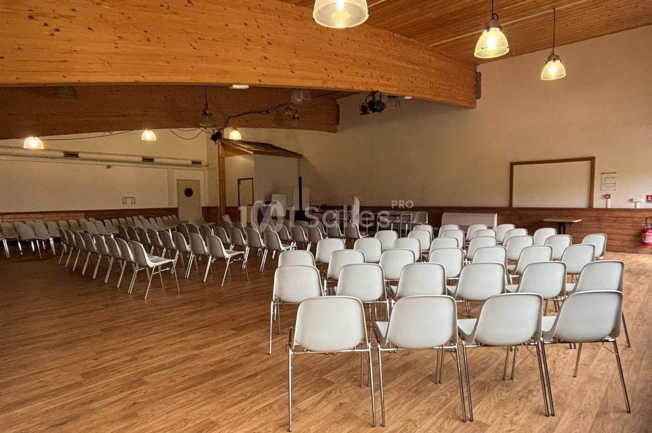 Salle avec parquet, rangées de chaises blanches alignées, éclairage suspendu et estrade en fond.