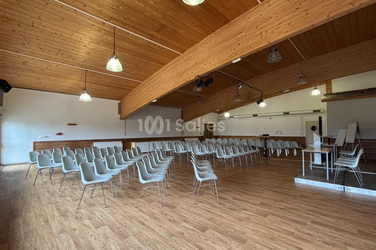 Salle de réunion vide avec des rangées de chaises blanches, parquet en bois et plafond avec poutres apparentes.