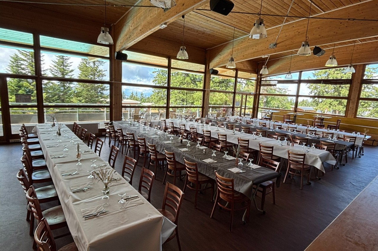Salle lumineuse avec de grandes baies vitrées, tables longues dressées avec nappes blanches et grises, chaises en bois.