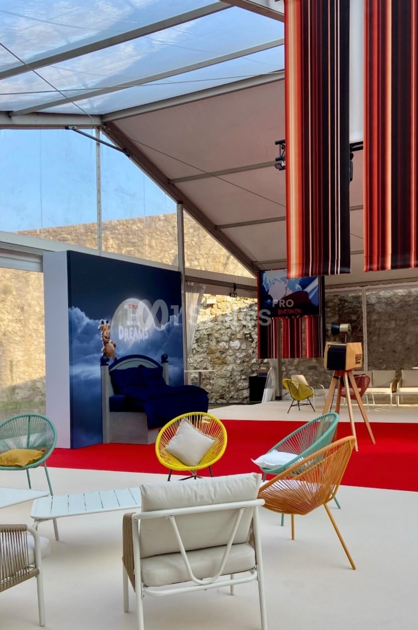 Espace lounge sous une structure transparente, avec fauteuils colorés, tapis rouge et décorations suspendues.