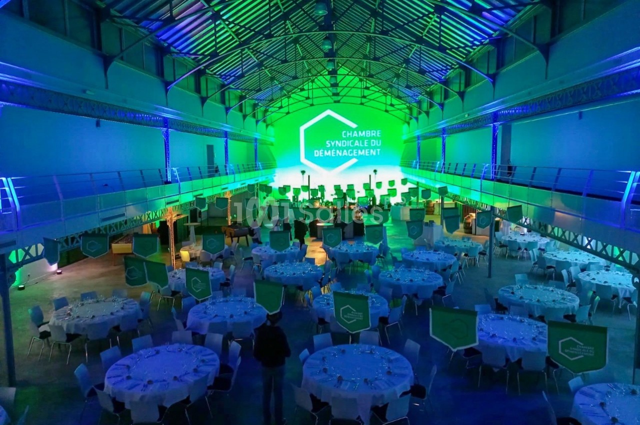 Salle de réception illuminée en vert avec des tables rondes dressées et une scène au fond pour un événement.