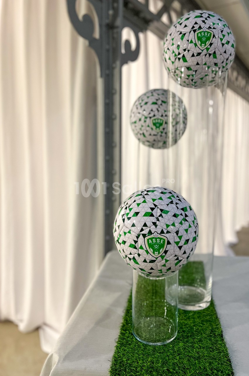 Trois ballons de football décoratifs aux motifs verts et blancs, posés sur des vases en verre sur une table.