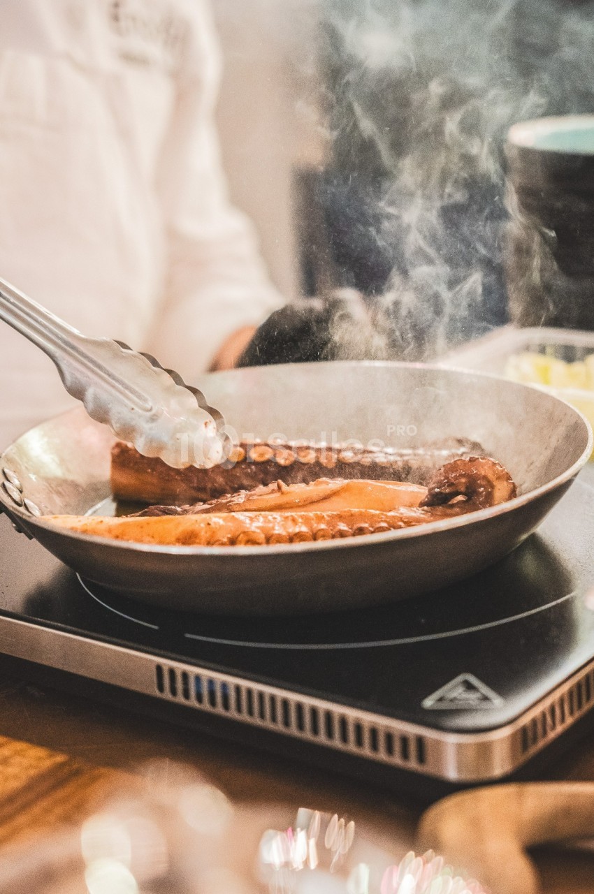 Tentacules de poulpe grillés dans une poêle chaude, manipulés avec une pince en cuisine.