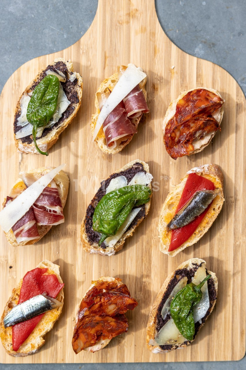 Assortiment de tapas sur une planche en bois, garnis de légumes, charcuterie, poissons et fromage.