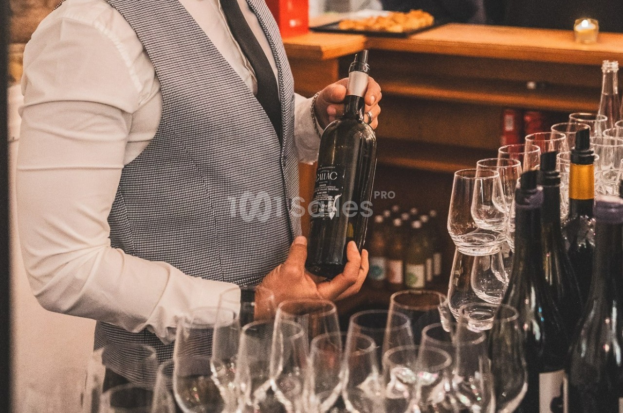 Un serveur en gilet présente une bouteille de vin derrière un comptoir avec des verres alignés.