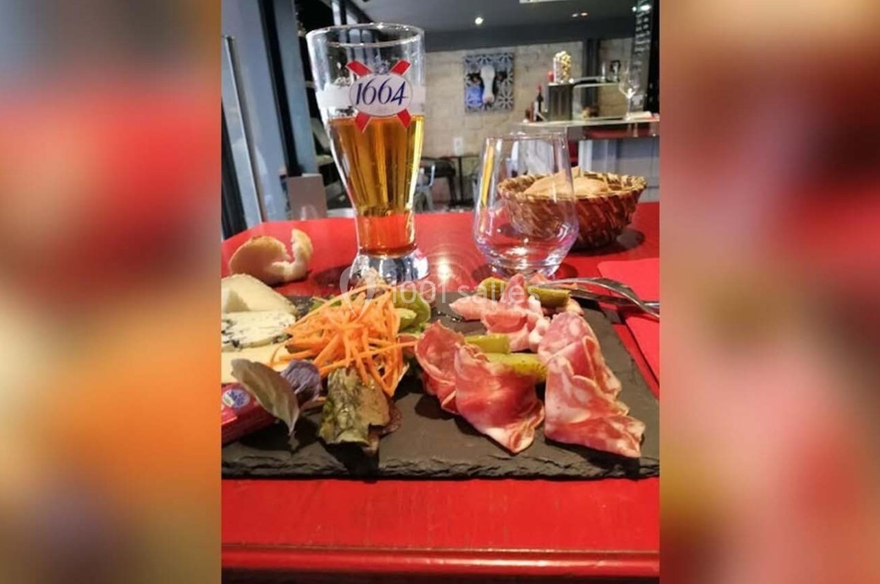 Assiette de charcuterie et fromages avec légumes, accompagnée d'une bière et d'un verre d'eau sur une table rouge.