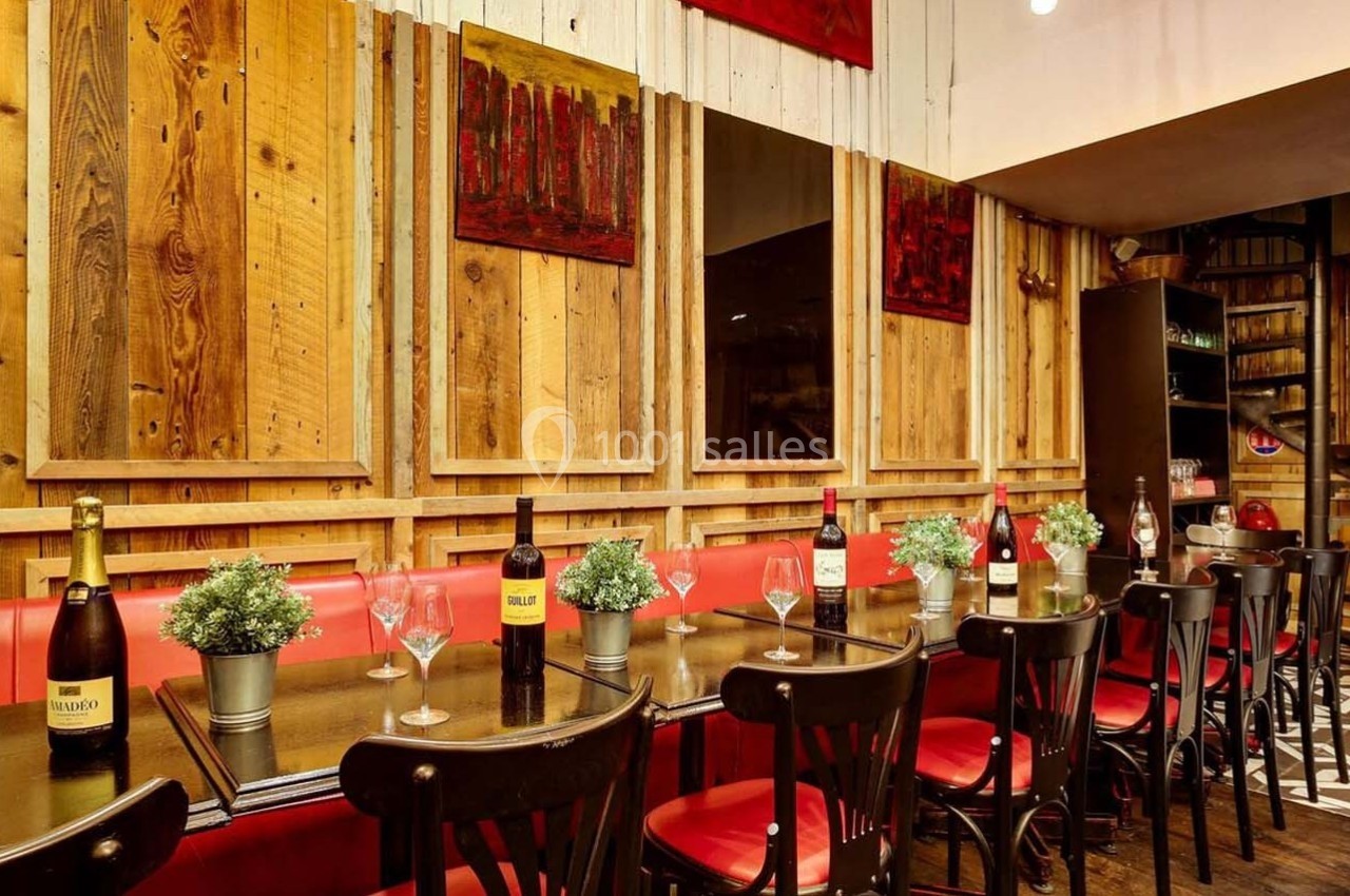 Salle de restaurant avec tables alignées, chaises noires, bouteilles de vin, plantes décoratives et murs en bois.