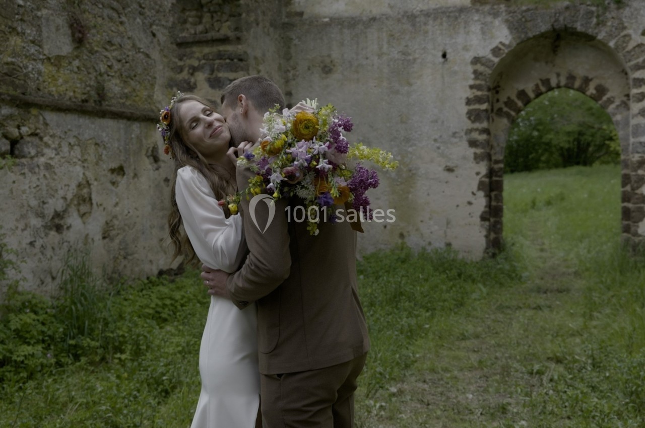 Un couple s'enlace tendrement devant un mur en pierre ancienne, dans un cadre verdoyant et fleuri.