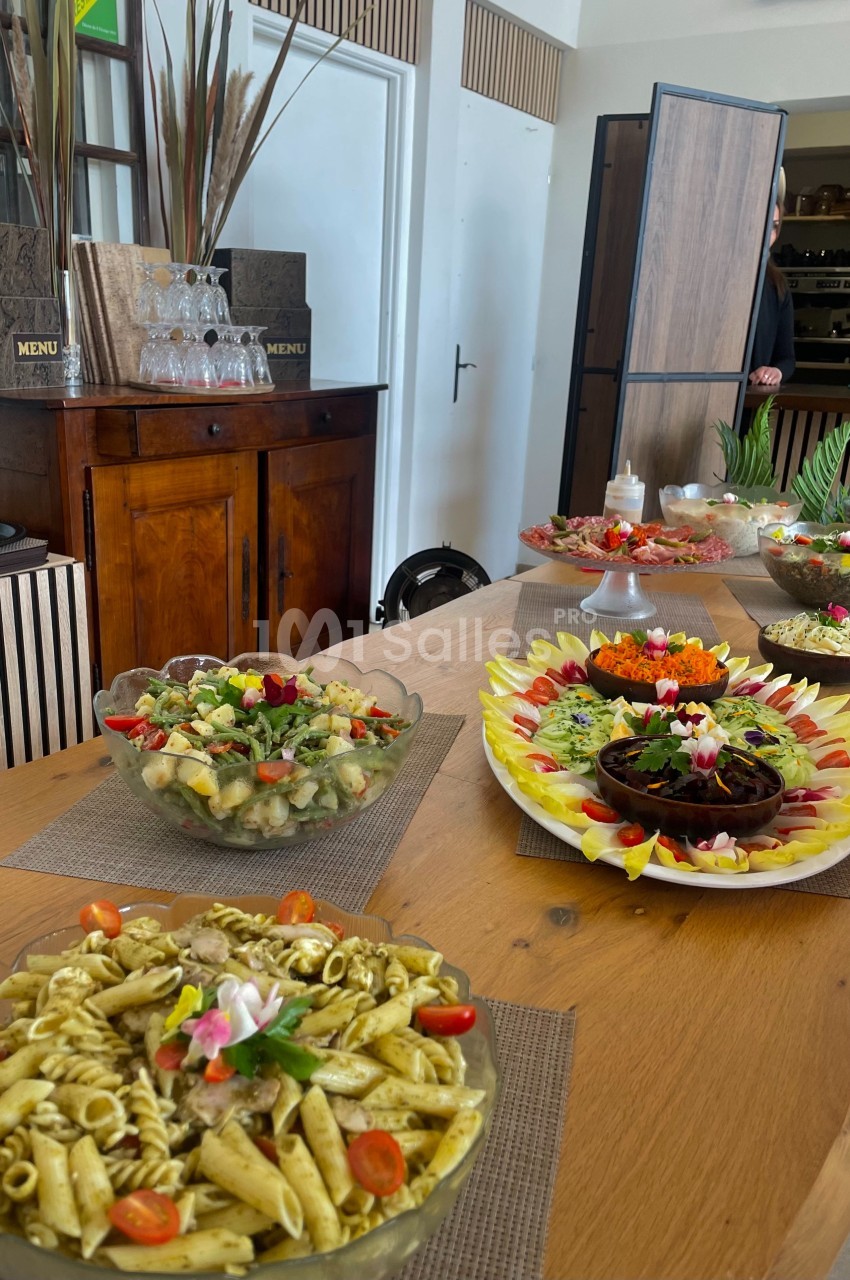 Buffet de plats variés comprenant des pâtes, salades et légumes, disposés sur une table en bois dans une salle lumineuse.