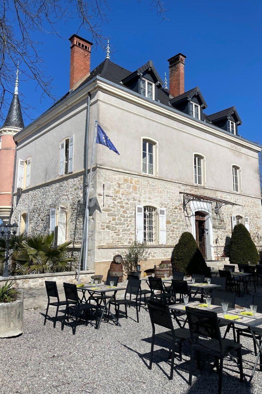 Façade d'un bâtiment ancien avec terrasse aménagée, tables et chaises, sous un ciel bleu dégagé.