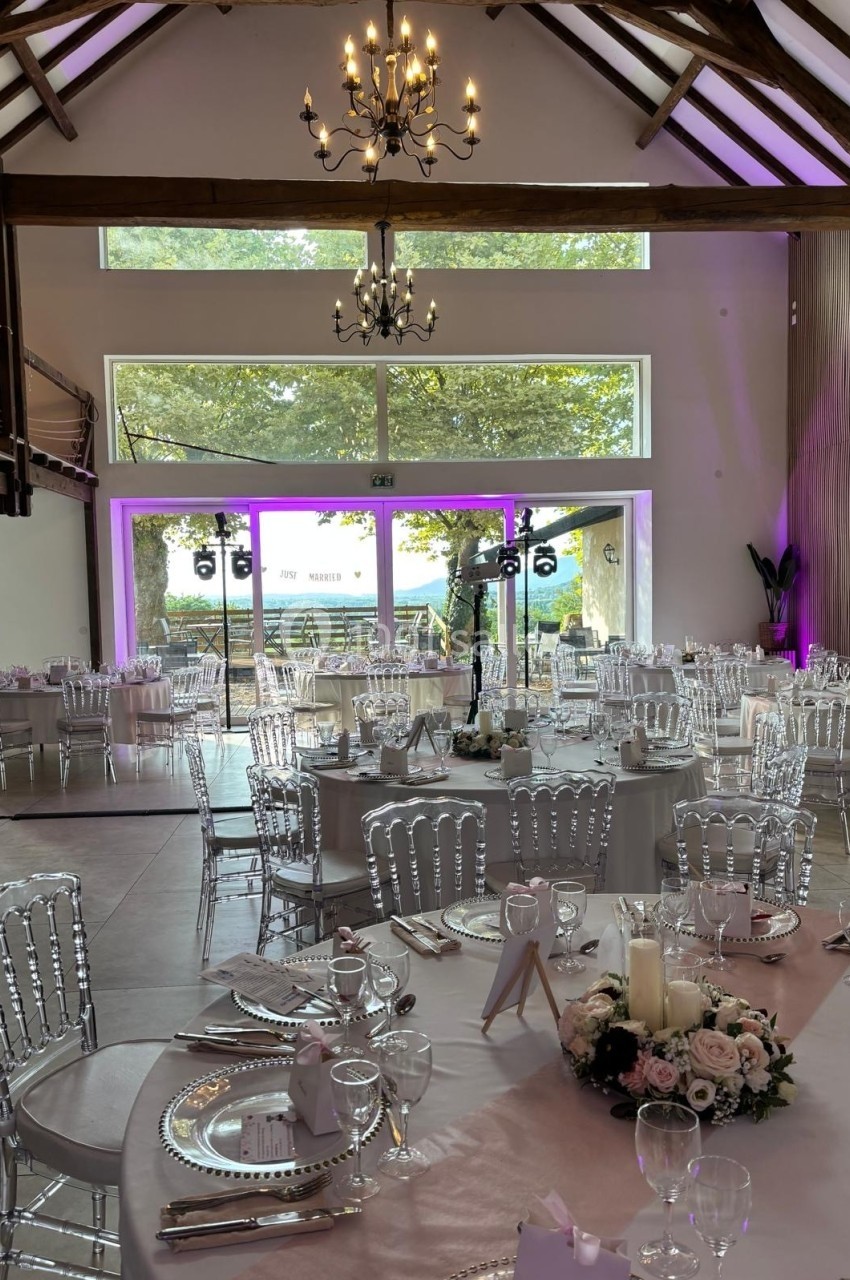 Salle de réception lumineuse avec tables décorées, chaises transparentes et vue sur un jardin à travers de grandes baies…