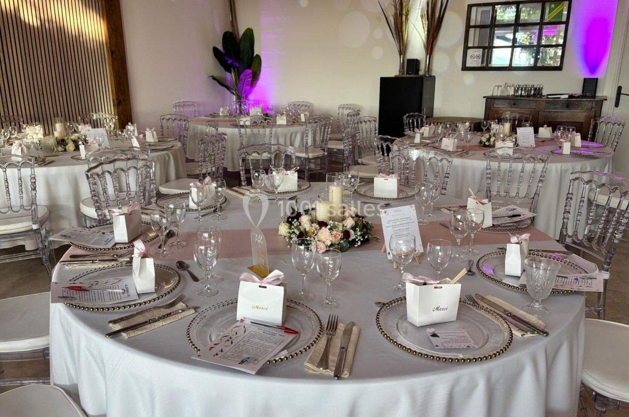 Salle de réception décorée avec des tables rondes dressées, nappes blanches, chaises transparentes et centre de table floral.