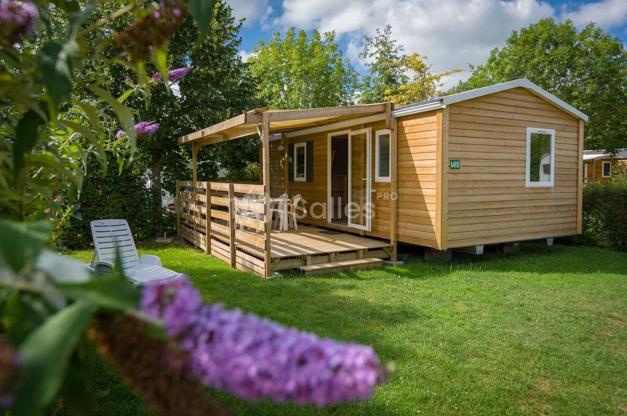 Chalet en bois avec terrasse couverte, entouré de verdure et fleurs violettes au premier plan.