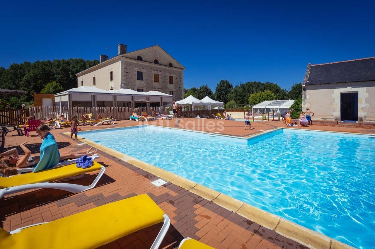 Piscine extérieure entourée de transats jaunes, située devant un bâtiment en pierre et des espaces ombragés.