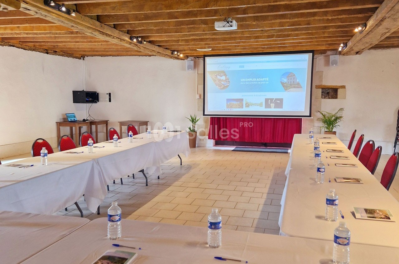 Salle de réunion lumineuse avec tables disposées en U, écran de projection, chaises rouges et poutres apparentes.
