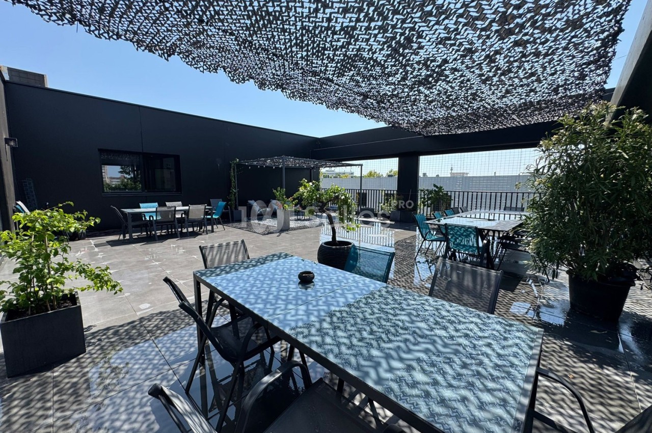 Terrasse extérieure avec tables, chaises, plantes en pots et ombrage en filet sous un ciel dégagé.