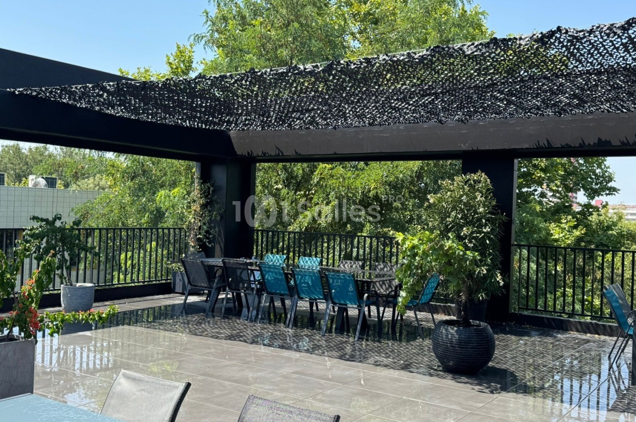 Terrasse extérieure avec tables, chaises bleues et noires, pergola en filet d'ombrage et végétation environnante.