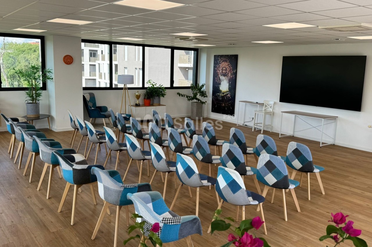 Salle lumineuse avec des rangées de chaises colorées, un écran noir au mur et des plantes en décoration.