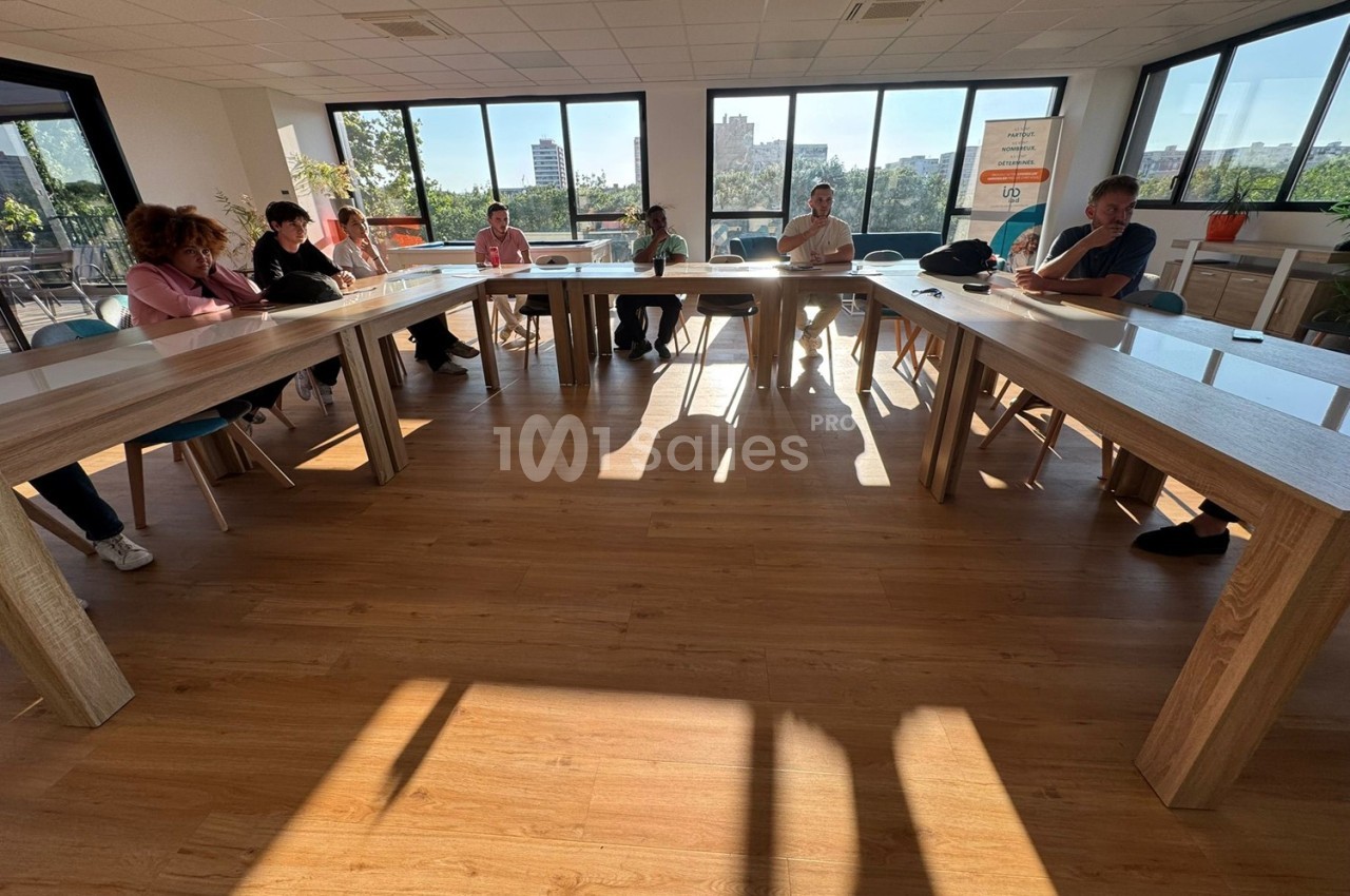 Personnes assises autour de tables en U dans une salle lumineuse avec de grandes fenêtres donnant sur l'extérieur.