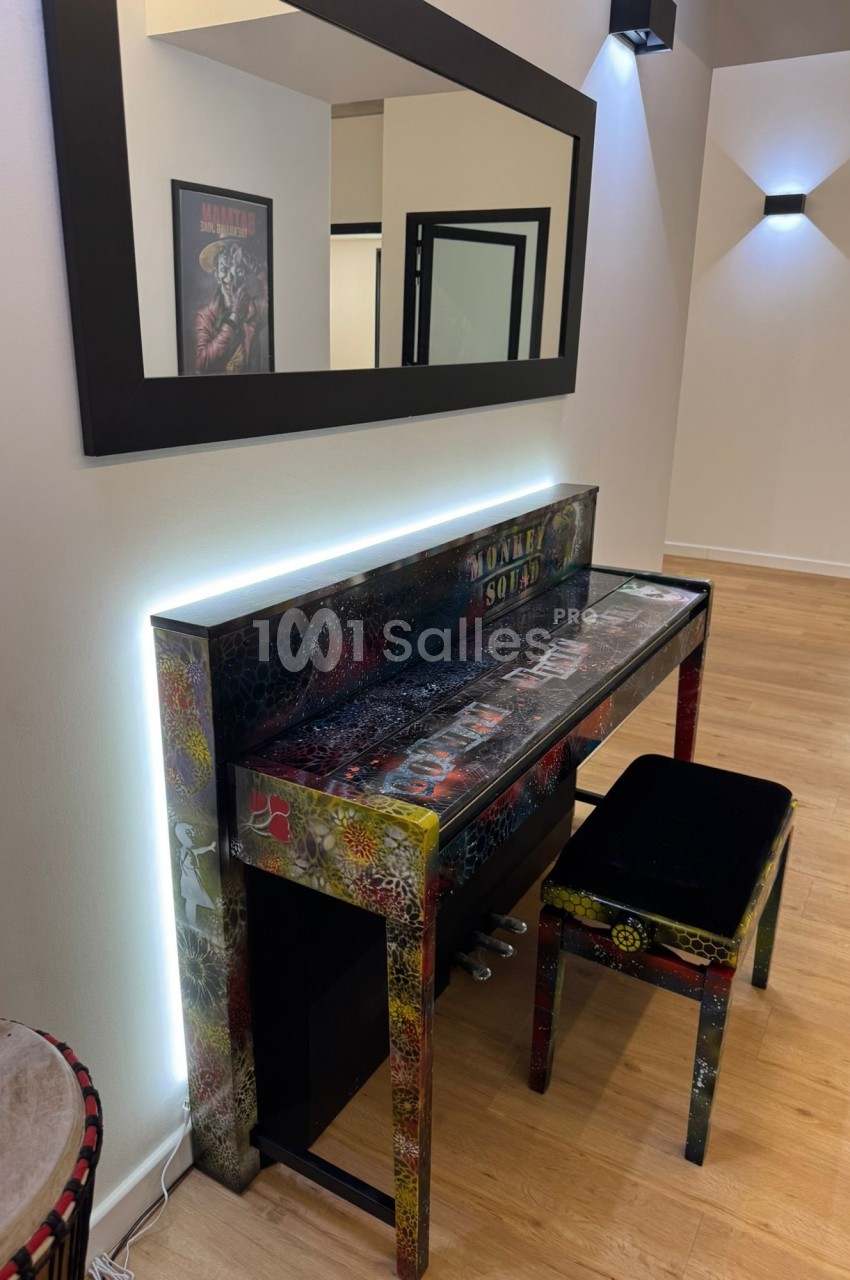 Piano droit coloré avec motifs artistiques, accompagné d'un tabouret noir, dans une pièce moderne au sol en bois.