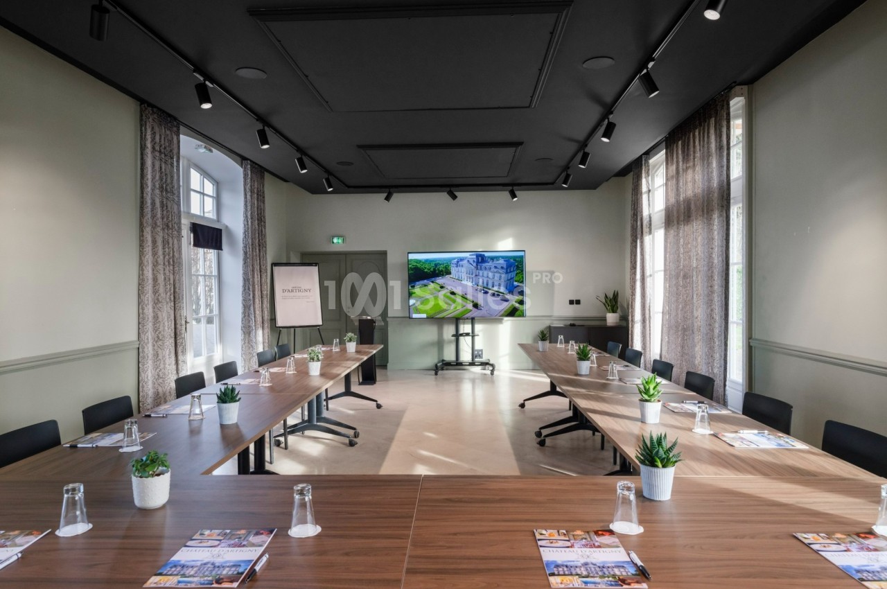 Salle de réunion lumineuse avec tables disposées en U, écran de présentation et plantes décoratives.