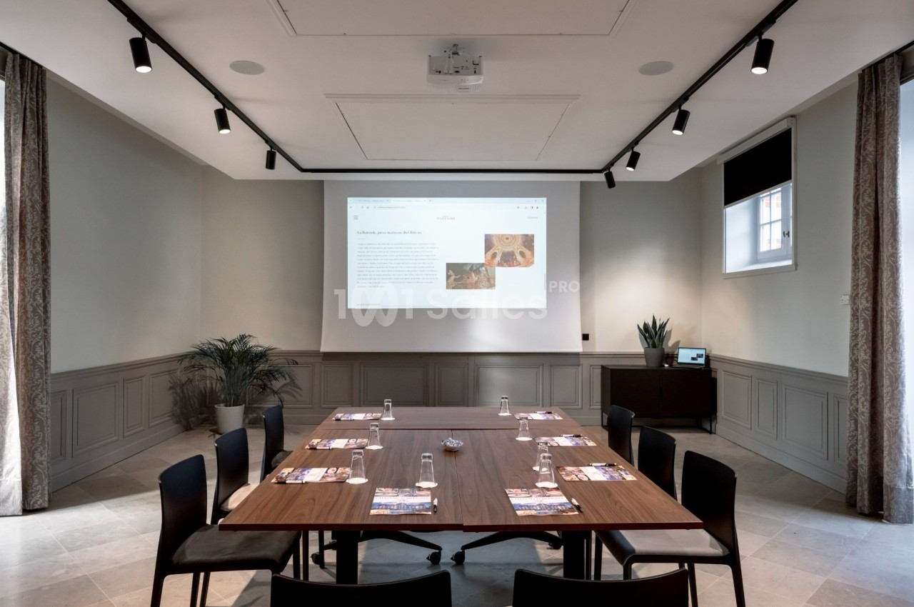 Salle de réunion lumineuse avec table en bois, chaises noires, écran de projection et plantes décoratives.