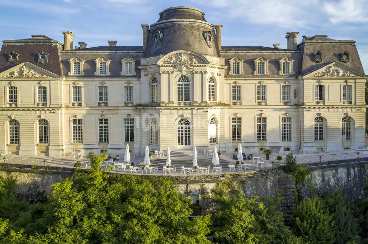 Façade d'un château historique avec terrasses et parasols, entouré de verdure sous un ciel dégagé.