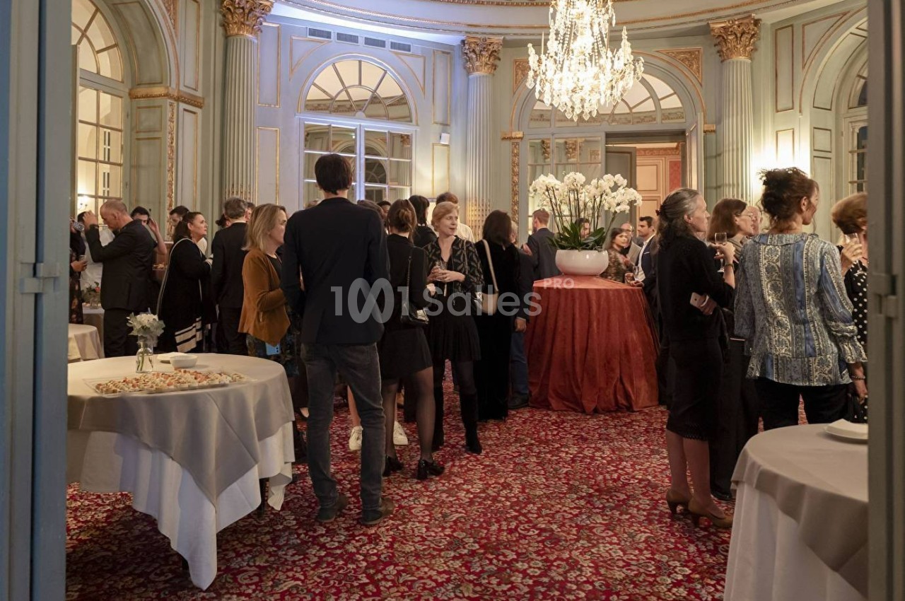 Groupe de personnes échangeant dans une salle élégante avec lustre, grandes fenêtres et tables décorées.
